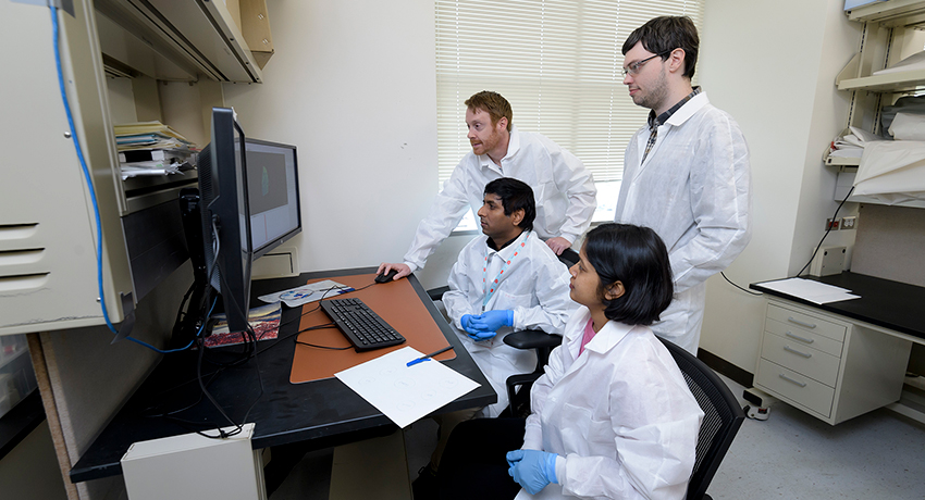 A professor in a white coat works on a computer in a laboratory while three students also in white lab coats observe.