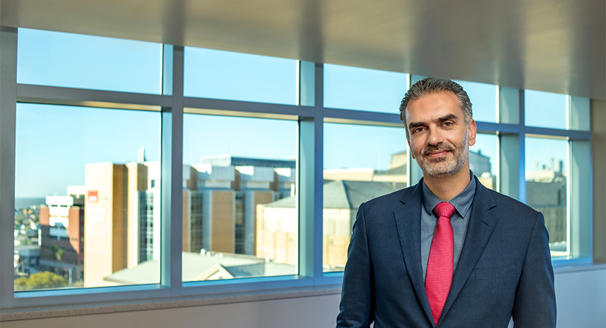 Person in a suit and red tie standing indoors with large windows showing a hospital in the background.