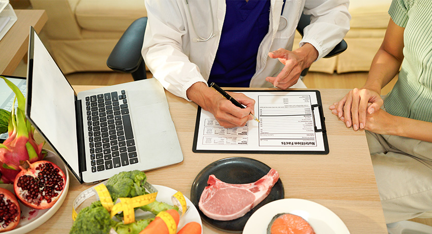 Doctor in a white coat reviews a nutrition plan on a clipboard with a patient at a desk; laptop open beside them, and various foods—fresh vegetables, pomegranate, salmon, and a raw pork chop—are arranged on the table, illustrating a dietary consultation.