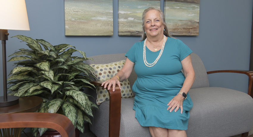 A woman in an aqua-colored dress smiles from her seat on the gray couch in her office.