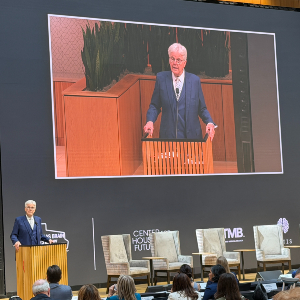  A speaker stands at a podium addressing an audience while a large screen above shows a live close-up of the same speaker; conference branding and empty stage chairs appear in the background. 