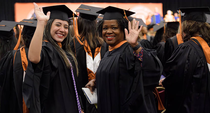 Two women in graduation regalia turn to wave as they prepare to receive their degrees.