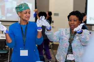 Two women in a training room wear gloves hold and spread white foam substance between their hands while standing at a table; presentation screen and other participants appear blurred in the background.