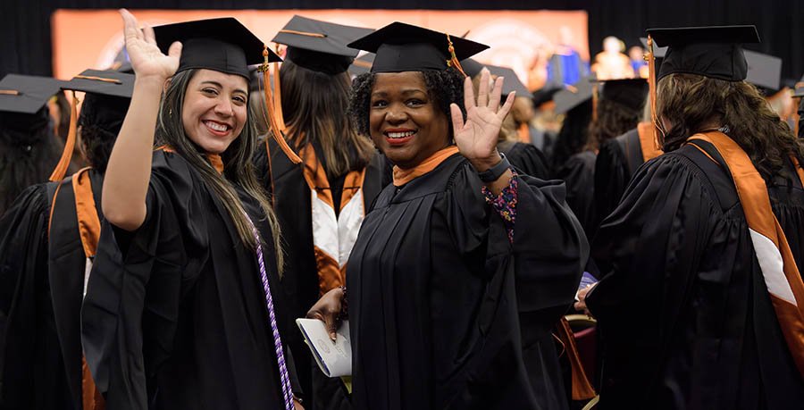 Two women in graduation regalia turn to wave as they prepare to receive their degrees.